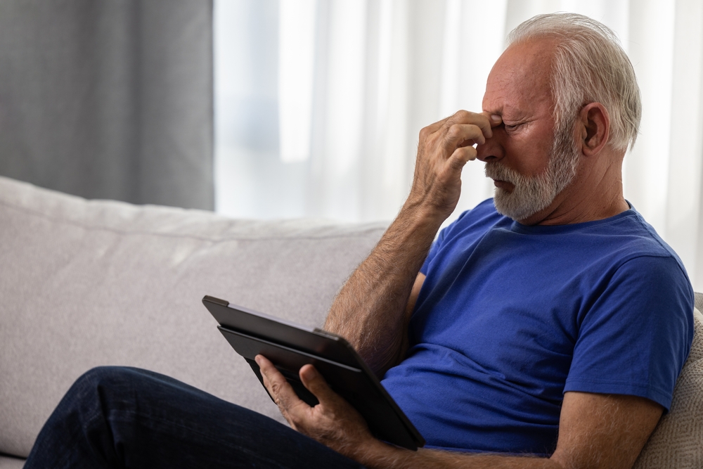 Elderly man rubbing eyes in frustration while using tablet on couch