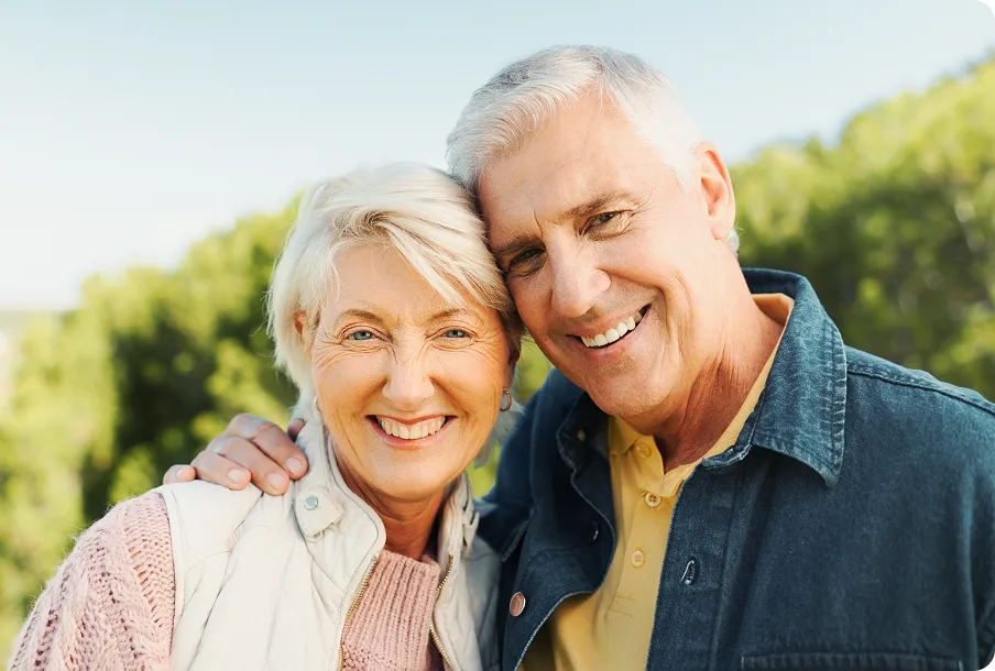 Happy elderly couple arm-in-arm in trees – nature adventures with clear post-surgery vision.